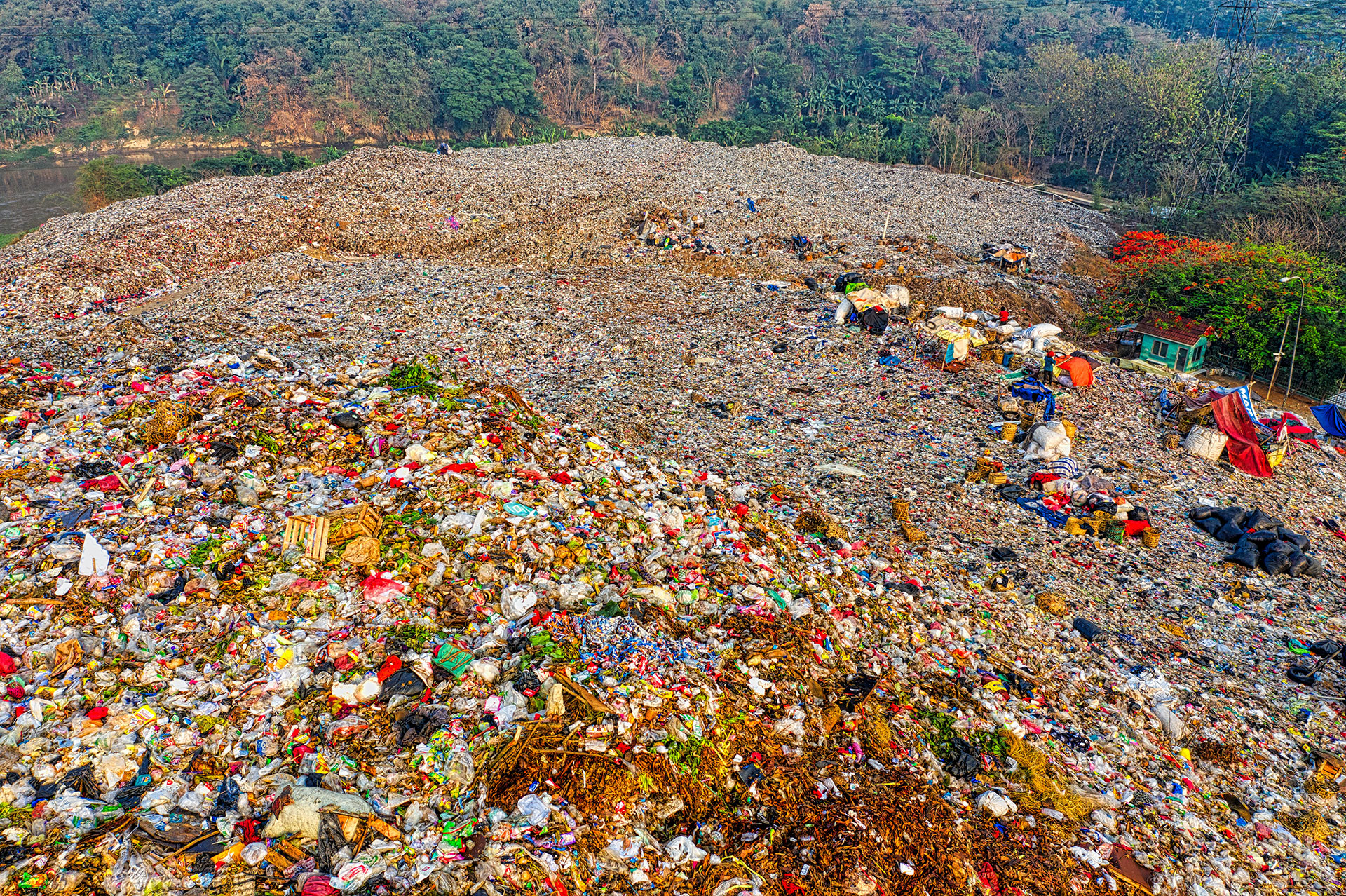 An image of a landfill spreading very far. Towards the bottom of the mountain of trash is a small green house and further behind that is a jungle forest.
